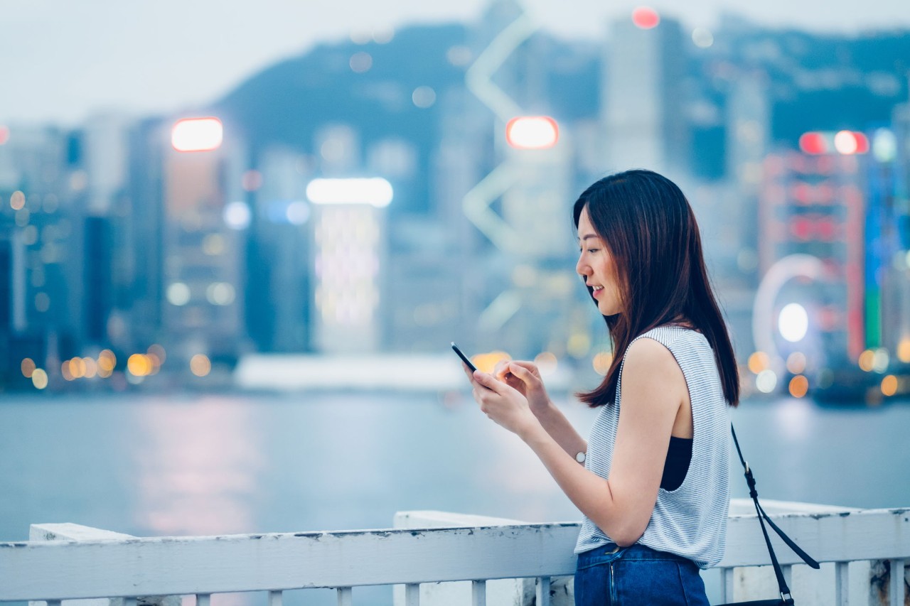 Smiling young lady using cell phone on urban rooftop, with illuminated city skyline as background